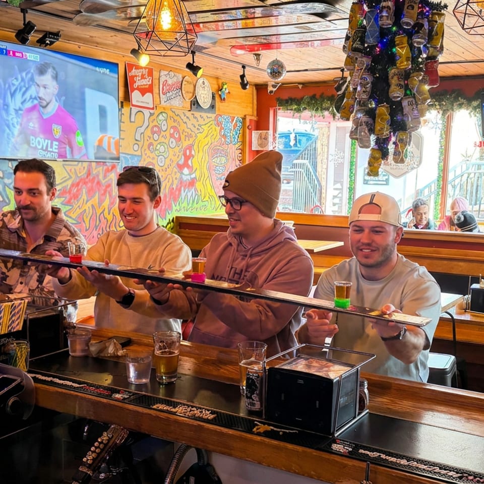 Friends in a colorful, graffiti-decorated bar hold a long serving tray while watching sports on television.