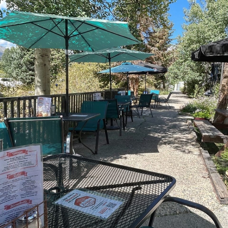 Outdoor restaurant patio with turquoise umbrellas, tables, menus, garden planters, and mountain views.