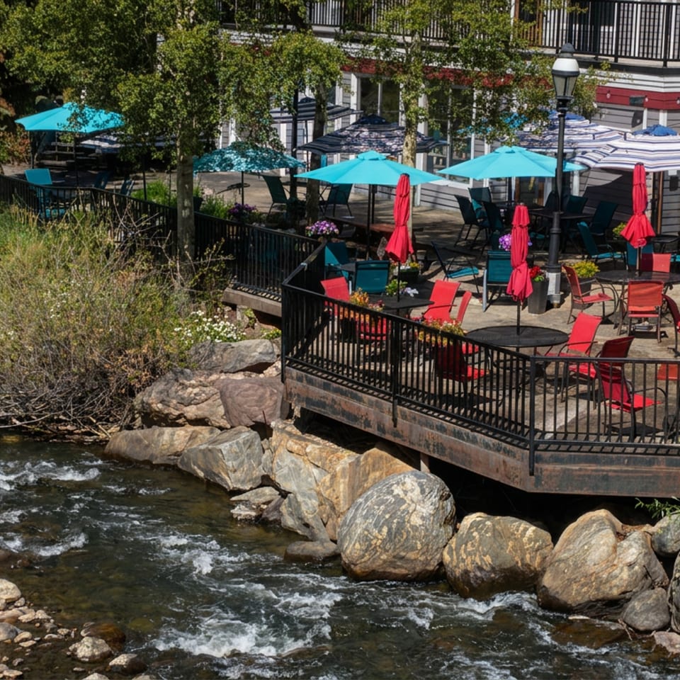 Riverside restaurant patio with red chairs, turquoise umbrellas, and tables overlooking a rocky, flowing stream.