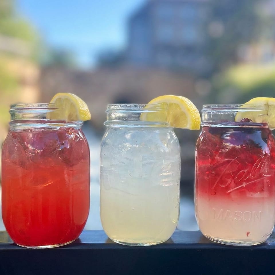 Three mason jars of colorful iced drinks garnished with lemon wedges, displayed outdoors against a softly blurred garden background.