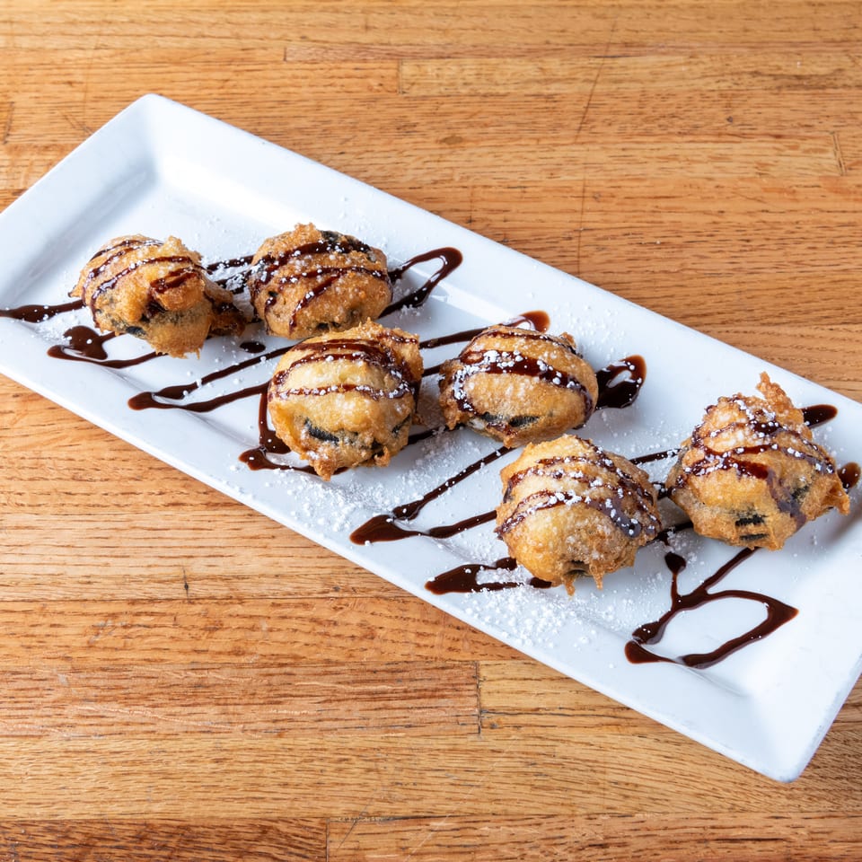 Six golden fried dessert bites on a white platter, drizzled with chocolate sauce and dusted with powdered sugar.
