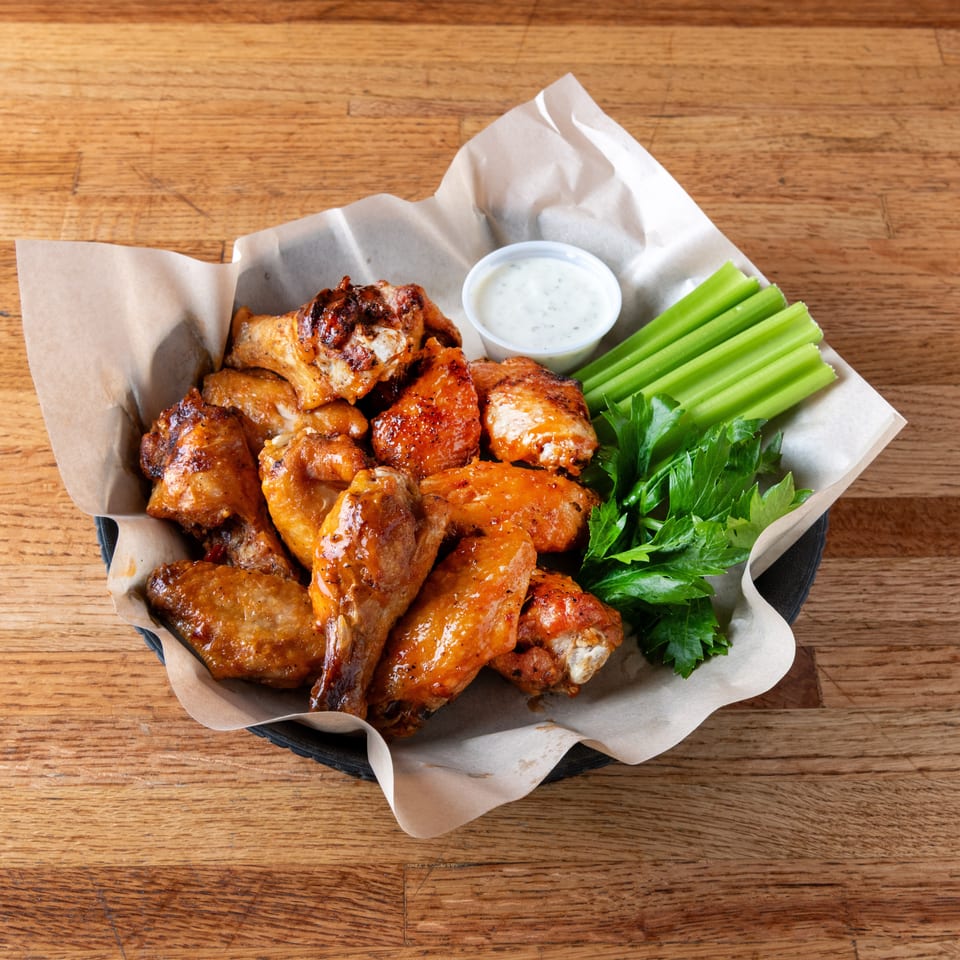 Basket of saucy chicken wings with celery sticks, ranch dipping sauce, and parsley on a wooden table.